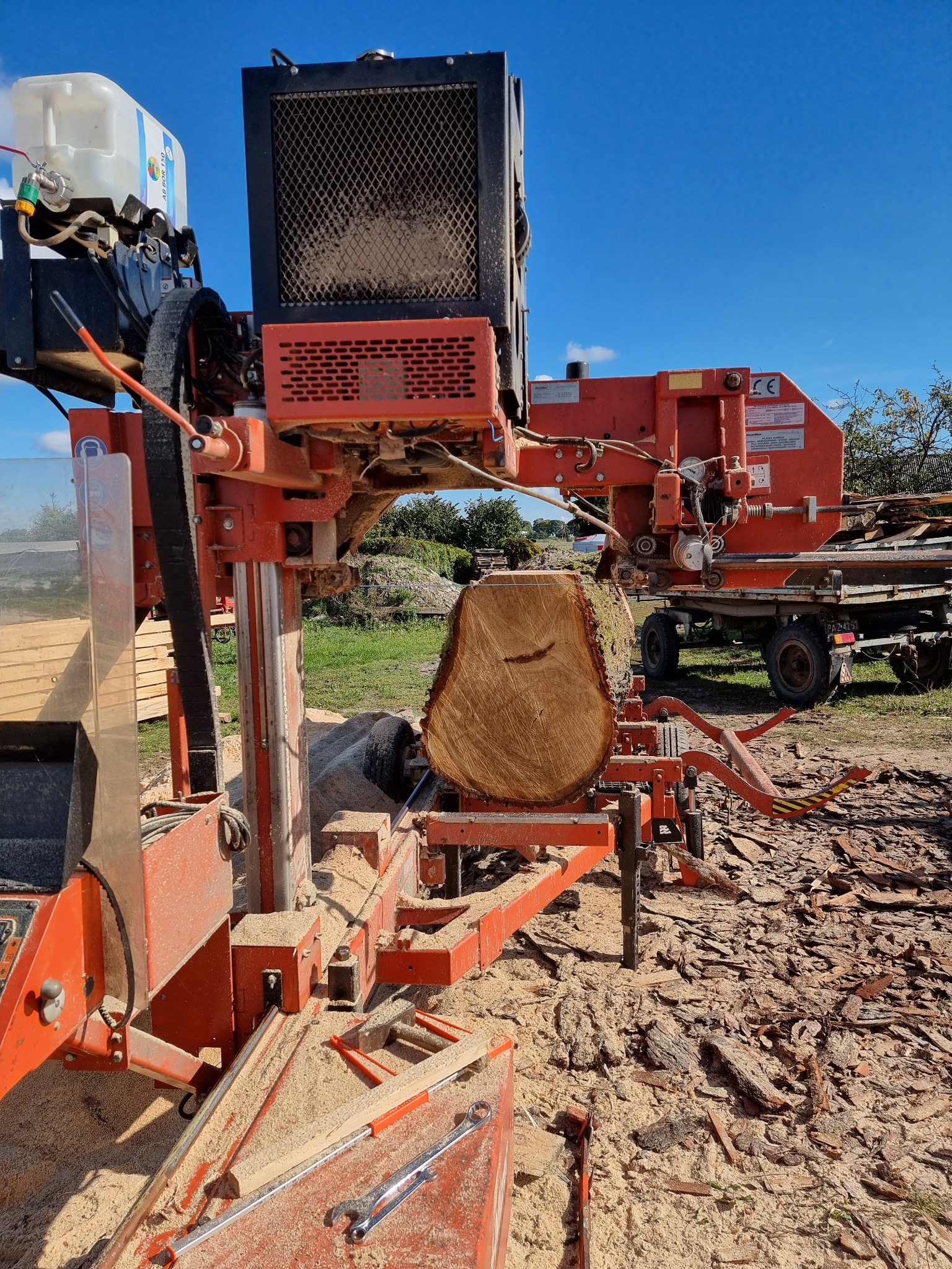 Proces cięcia kłody drewna na przenośnym traku taśmowym w plenerze, wióry i kora na ziemi, widoczne narzędzia