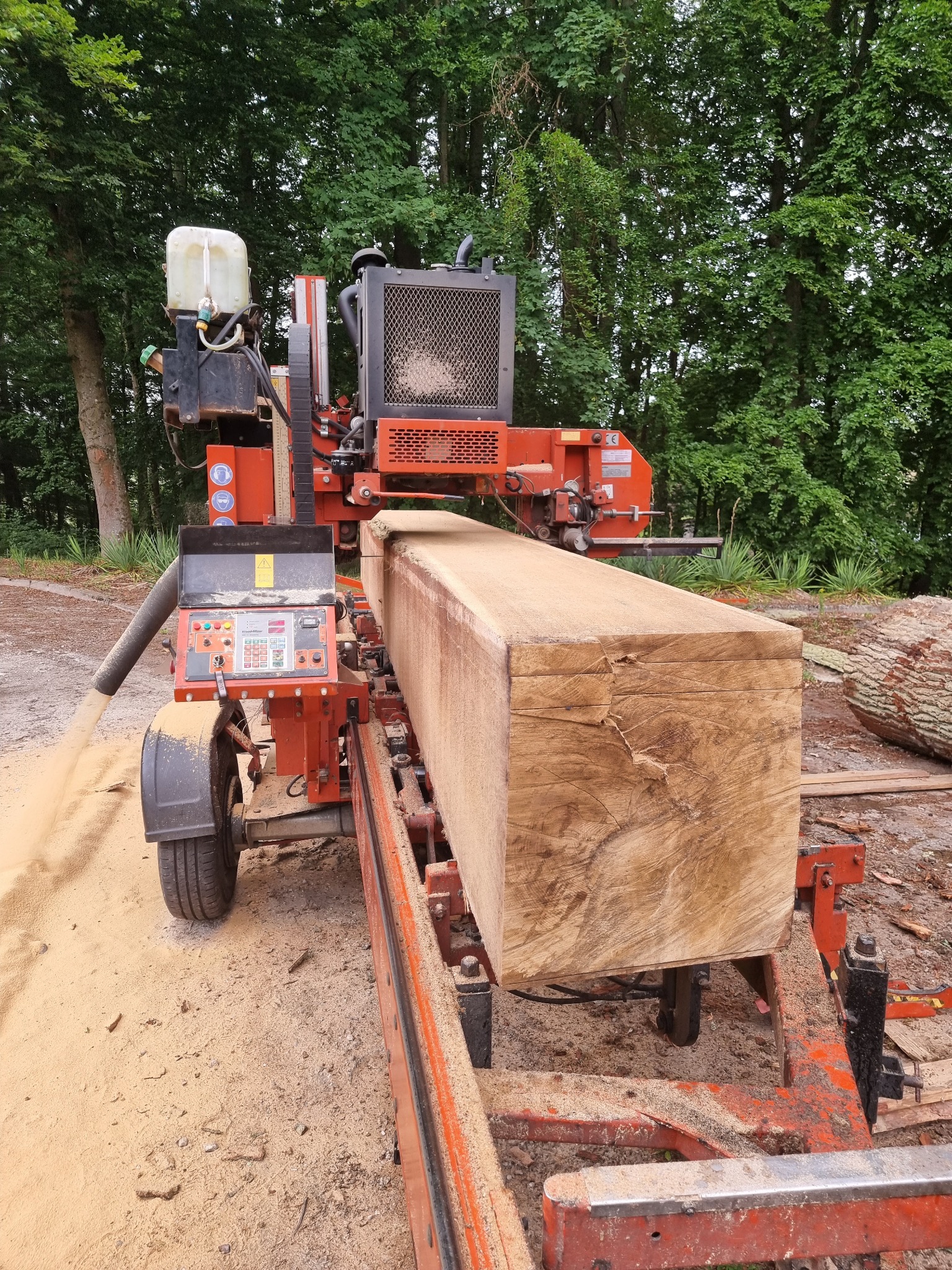 Mobilny tartak tnie kłodę drewna na deski, w tle zielone drzewa.