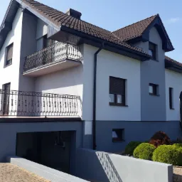 Dwukondygnacyjny dom z balkonami i garażem w piwnicy, wykończony w odcieniach szarości i bieli, z brązową dachówką i metalowymi balustradami balkonów o ozdobnym wzorze.