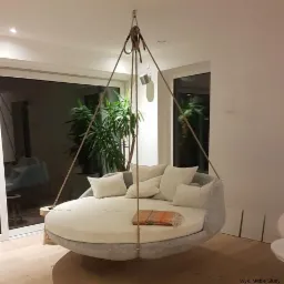 Nowoczesny salon z wiszącą, okrągłą sofą-bujawką podwieszoną na linach, z licznymi poduszkami i pledem, widok na taras z zielenią.