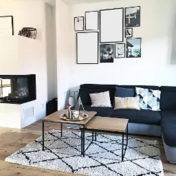 Nowoczesny salon z czarną sofą narożną, dywanem w geometryczne wzory, zestawem dwóch stolików kawowych z drewnianymi blatami i galerią plakatów w czarnych ramkach na białej ścianie.