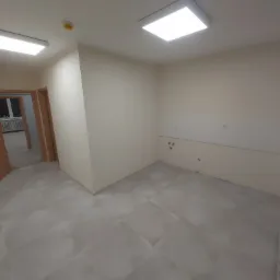 Pusta, świeżo wyremontowana łazienka z beżowymi ścianami, jasnymi płytkami podłogowymi i białymi płytkami na ścianie nad instalacją wodno-kanalizacyjną. Widoczne dwa otwory drzwiowe. W jednym...