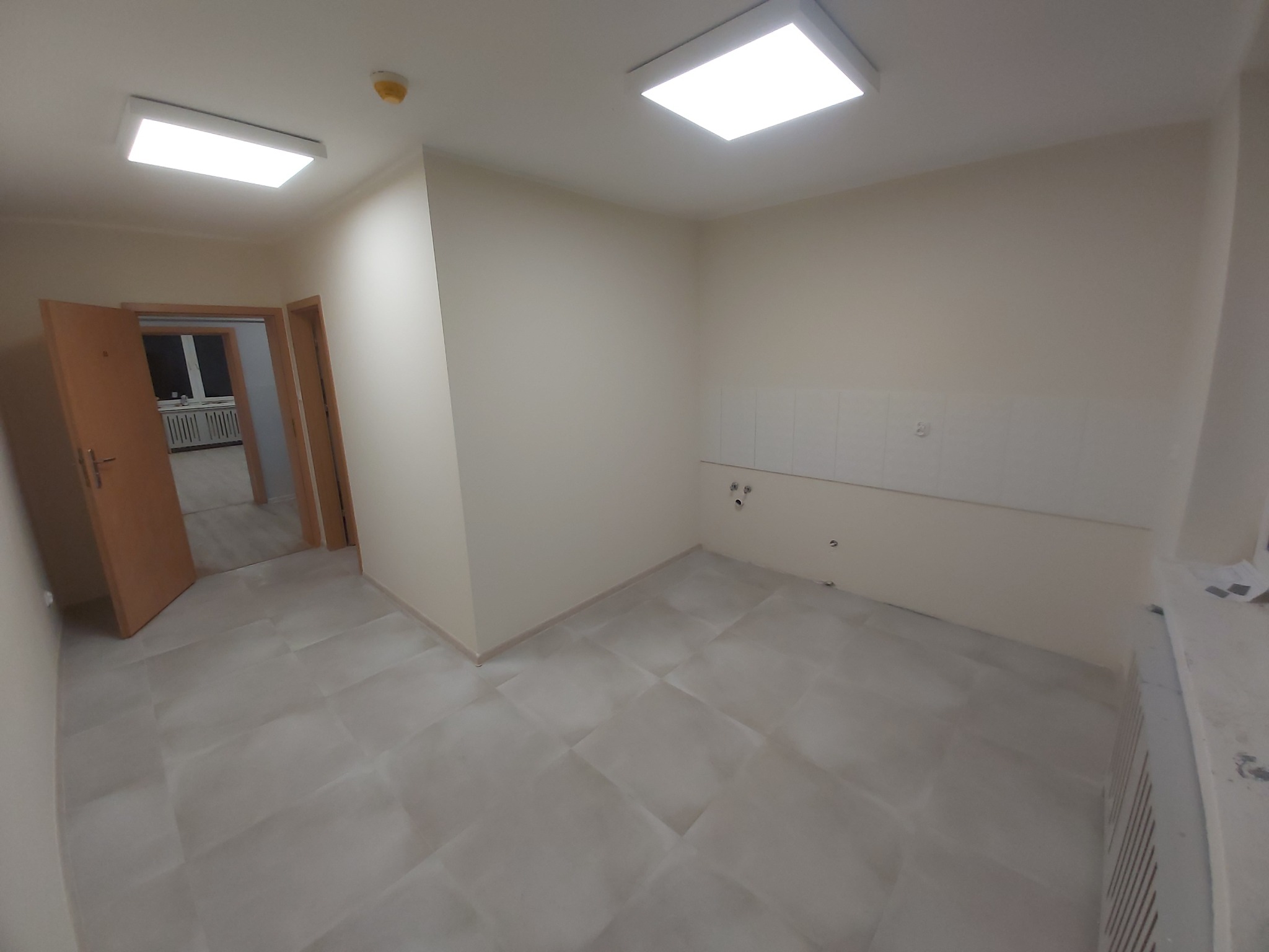 Pusta, świeżo wyremontowana łazienka z beżowymi ścianami, jasnymi płytkami podłogowymi i białymi płytkami na ścianie nad instalacją wodno-kanalizacyjną. Widoczne dwa otwory drzwiowe. W jednym...