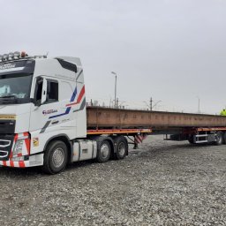 Auto-Matunin - Ciężarówka Volvo FH z naczepą przewożąca stalową belkę na tle torów kolejowych i żwirowego placu, widoczny pracownik w żółtej kamizelce.