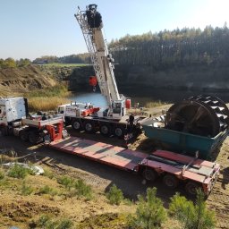 Auto-Matunin - Ciężarówka z naczepą niskopodwoziową oraz dźwig samochodowy na tle wyrobiska z wodą, transportujące element maszyny roboczej w kształcie koła łopatkowego.