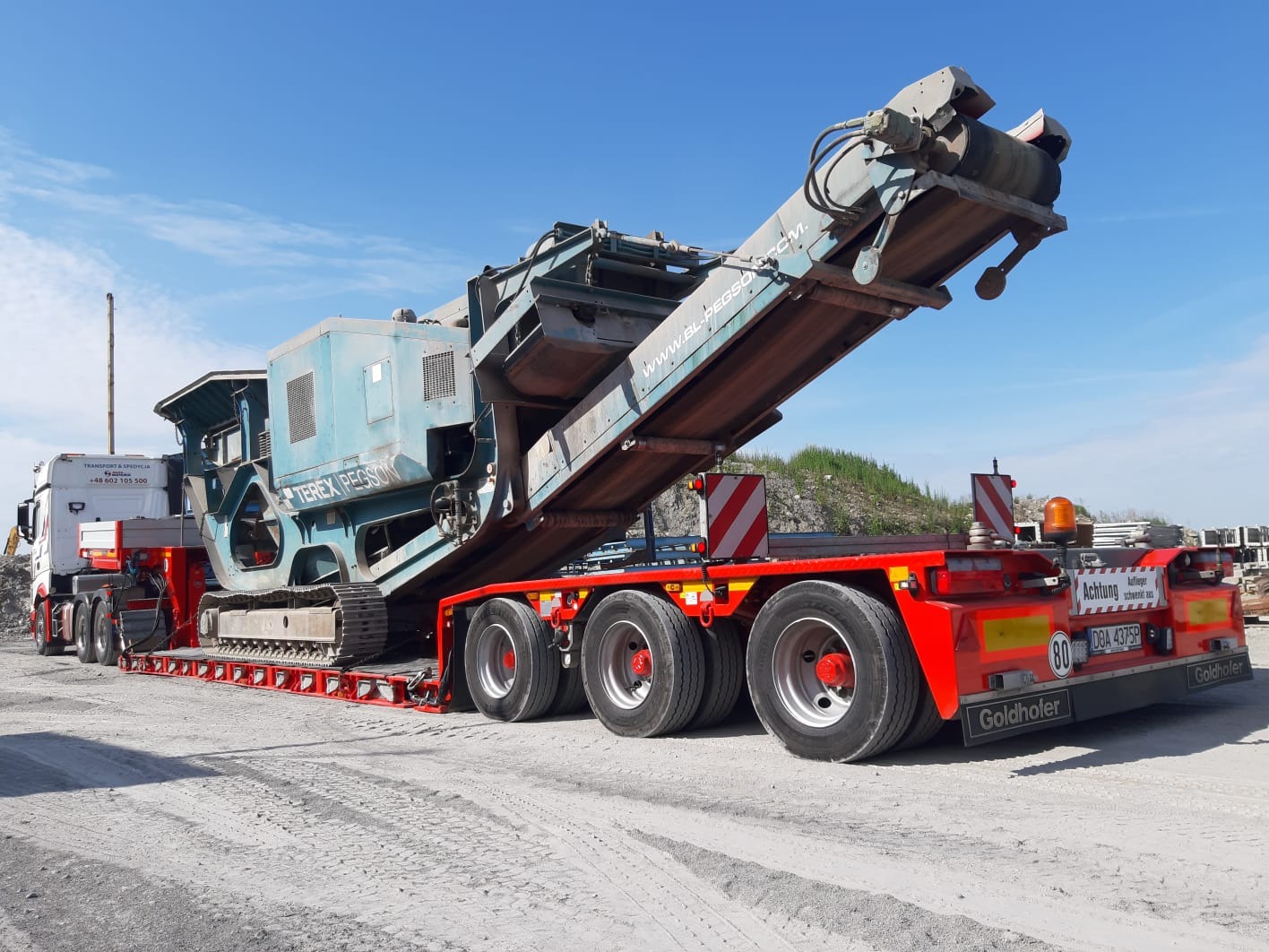 Ciężki, niebieski kruszarka Terex Pegson na czerwonej naczepie Goldhofer, przewożona przez białą ciężarówkę, widok z boku na tle błękitnego nieba.