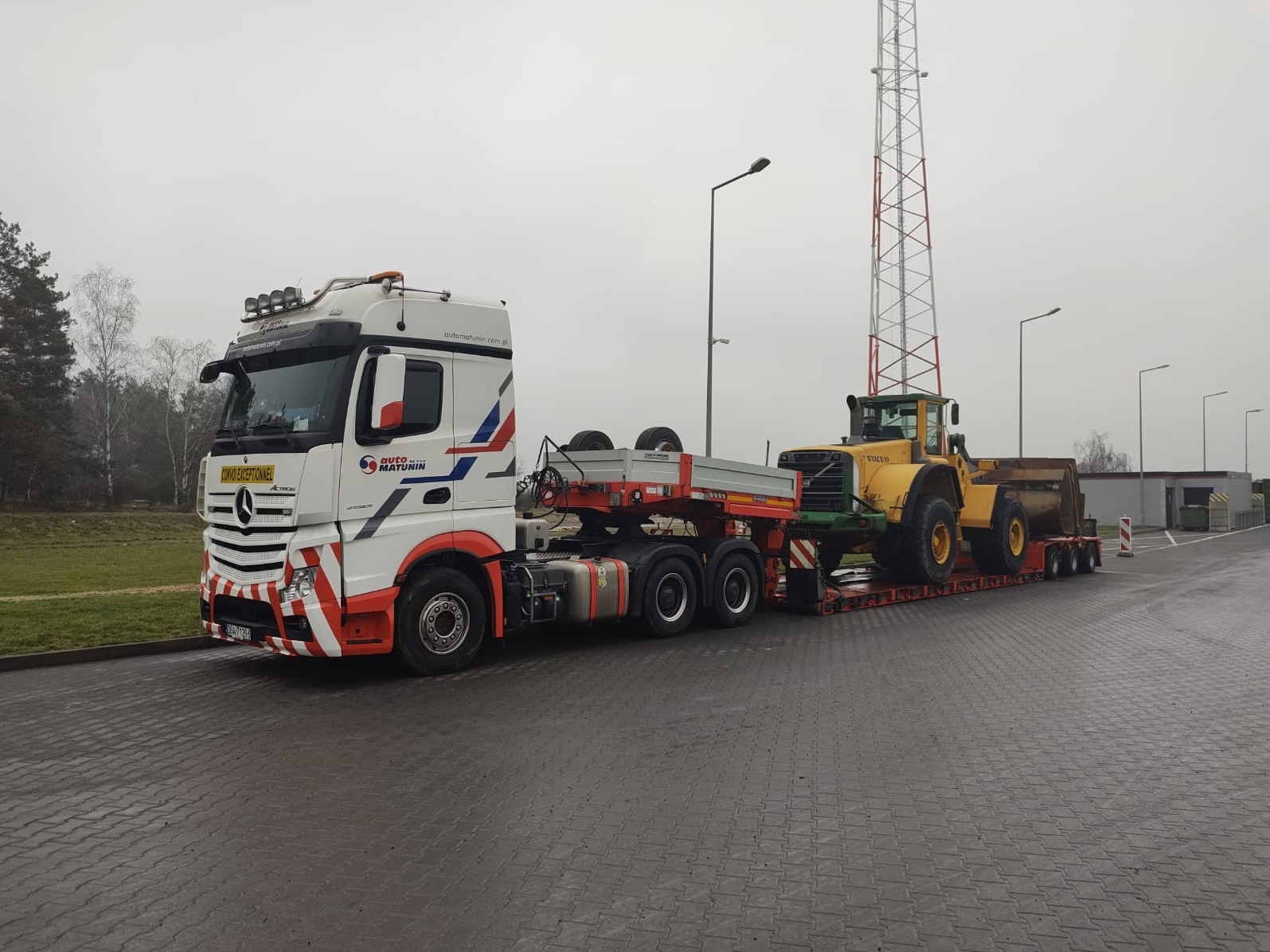 Ciężarówka Mercedes Actros z naczepą niskopodwoziową przewożąca żółtą ładowarkę Volvo na mokrej nawierzchni, widoczna wieża telekomunikacyjna w tle.