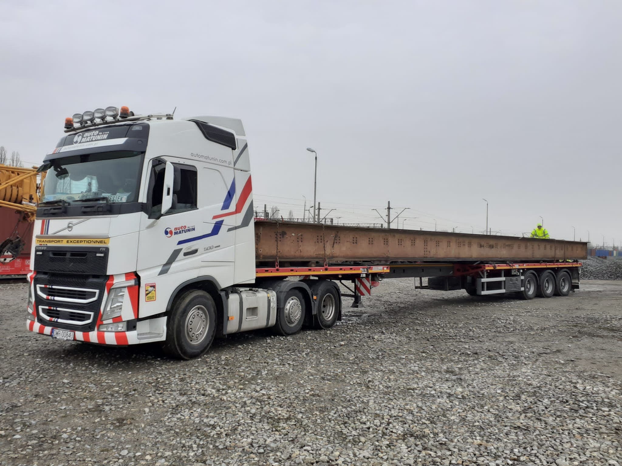 Ciężarówka Volvo FH z naczepą przewożąca stalową belkę na tle torów kolejowych i żwirowego placu, widoczny pracownik w żółtej kamizelce.