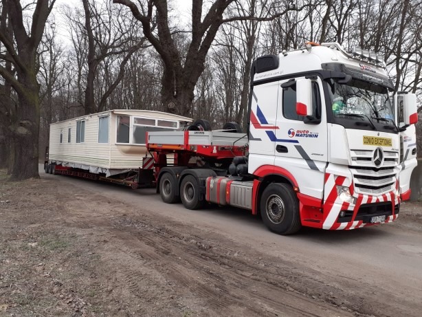 Ciężarówka marki Mercedes-Benz z naczepą niskopodwoziową przewożąca modułowy dom w kremowym kolorze, widoczny napis 'CONVOI EXCEPTIONNEL' na kabinie