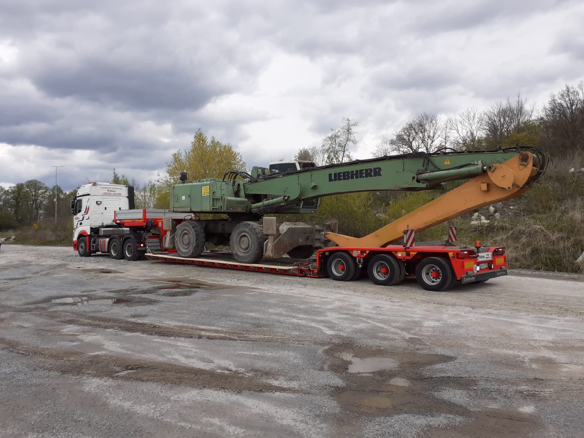 Transport ciężkiego sprzętu: Zielony dźwig Liebherr na czerwonej platformie transportowej, ciągnięty przez białą ciężarówkę, na szutrowym placu w pochmurny dzień.