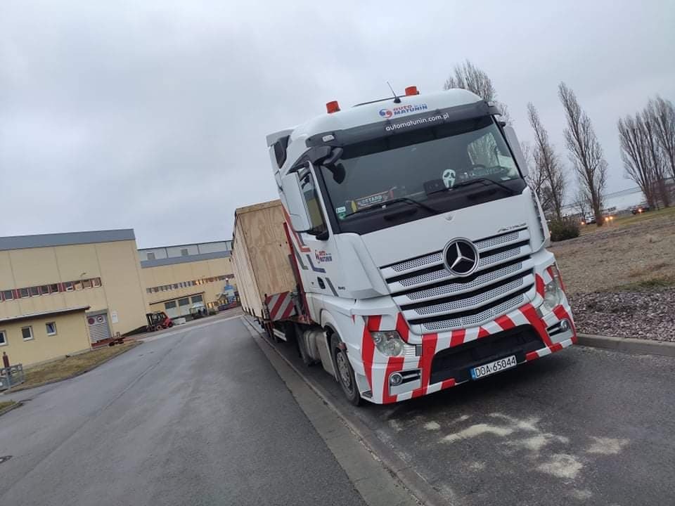 Biała ciężarówka Mercedes Actros z ładunkiem w drewnianej skrzyni, zaparkowana na poboczu drogi w pochmurny dzień. Na przedniej szybie widoczna naklejka 'Retarder'.