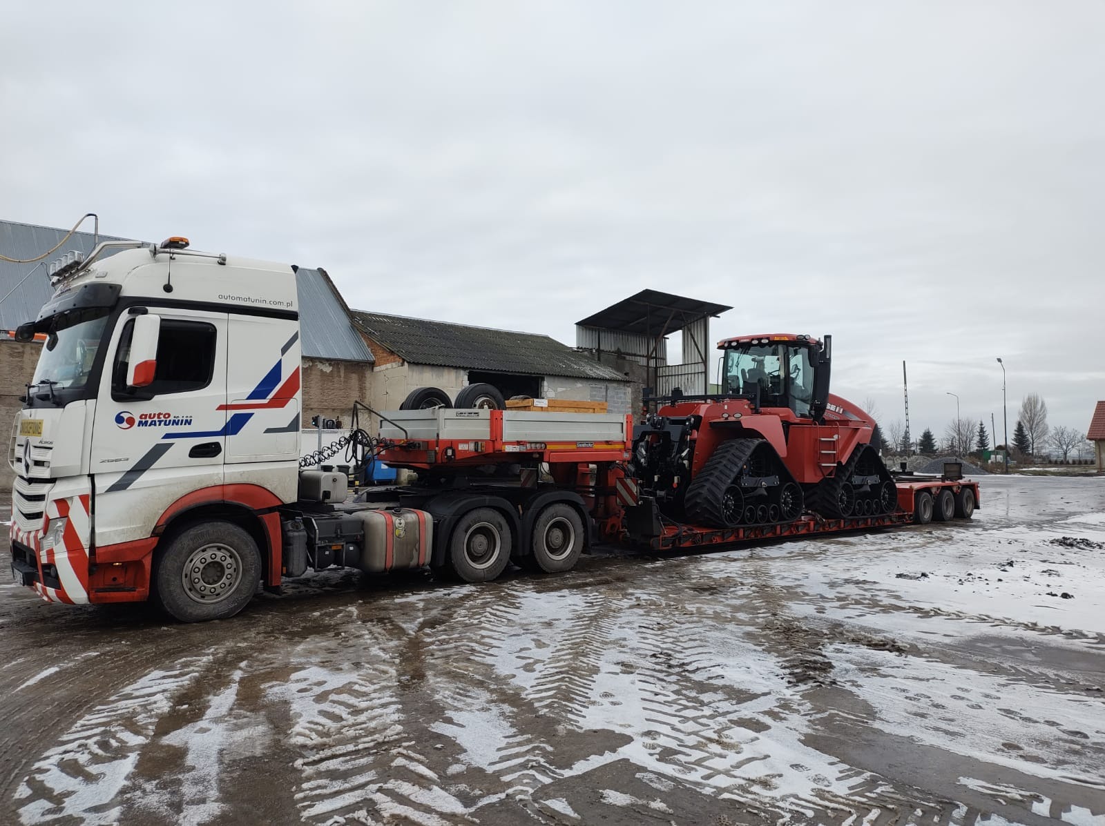 Czerwony ciągnik gąsienicowy przewożony na niskopodwoziowej platformie przez białą ciężarówkę z logo firmy Auto Matunin, na tle szarego, pochmurnego nieba i zabudowań gospodarczych.