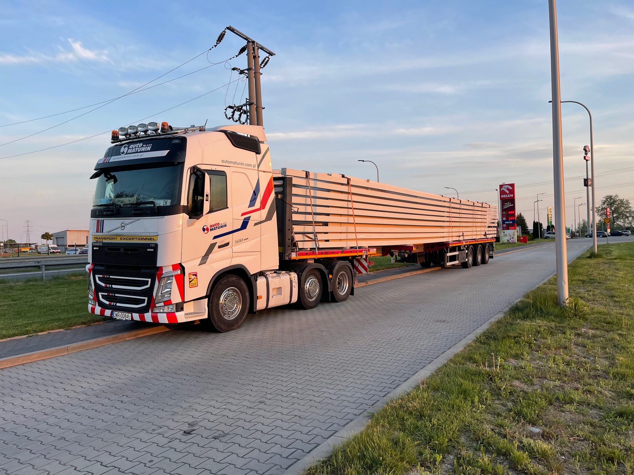 Ciężarówka Volvo FH 500 z naczepą, załadowana długimi, jasnymi profilami metalowymi, zaparkowana na parkingu przy stacji Orlen, widoczny słup wysokiego napięcia i niebieskie niebo