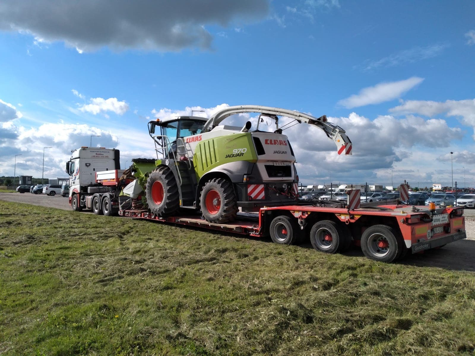Ciężarówka z naczepą niskopodwoziową przewożąca kombajn rolniczy Claas Jaguar 970 na tle parkingu samochodów osobowych i błękitnego nieba.