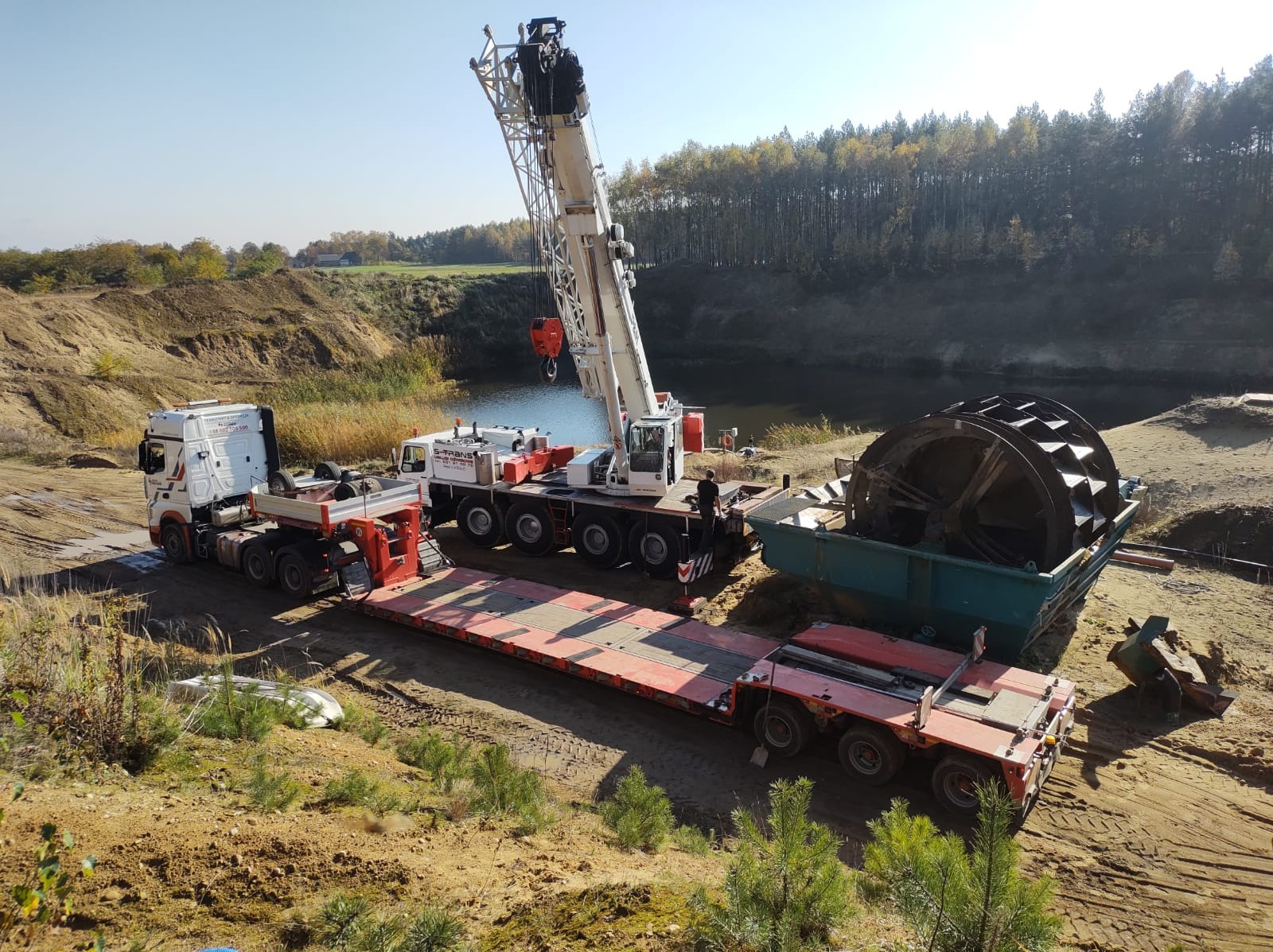 Ciężarówka z naczepą niskopodwoziową oraz dźwig samochodowy na tle wyrobiska z wodą, transportujące element maszyny roboczej w kształcie koła łopatkowego.