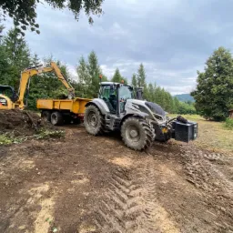 Żółta koparka załadowuje ziemię i gałęzie do przyczepy ciągniętej przez szary traktor na nierównym, błotnistym terenie, z widocznymi śladami opon i zielenią w tle.