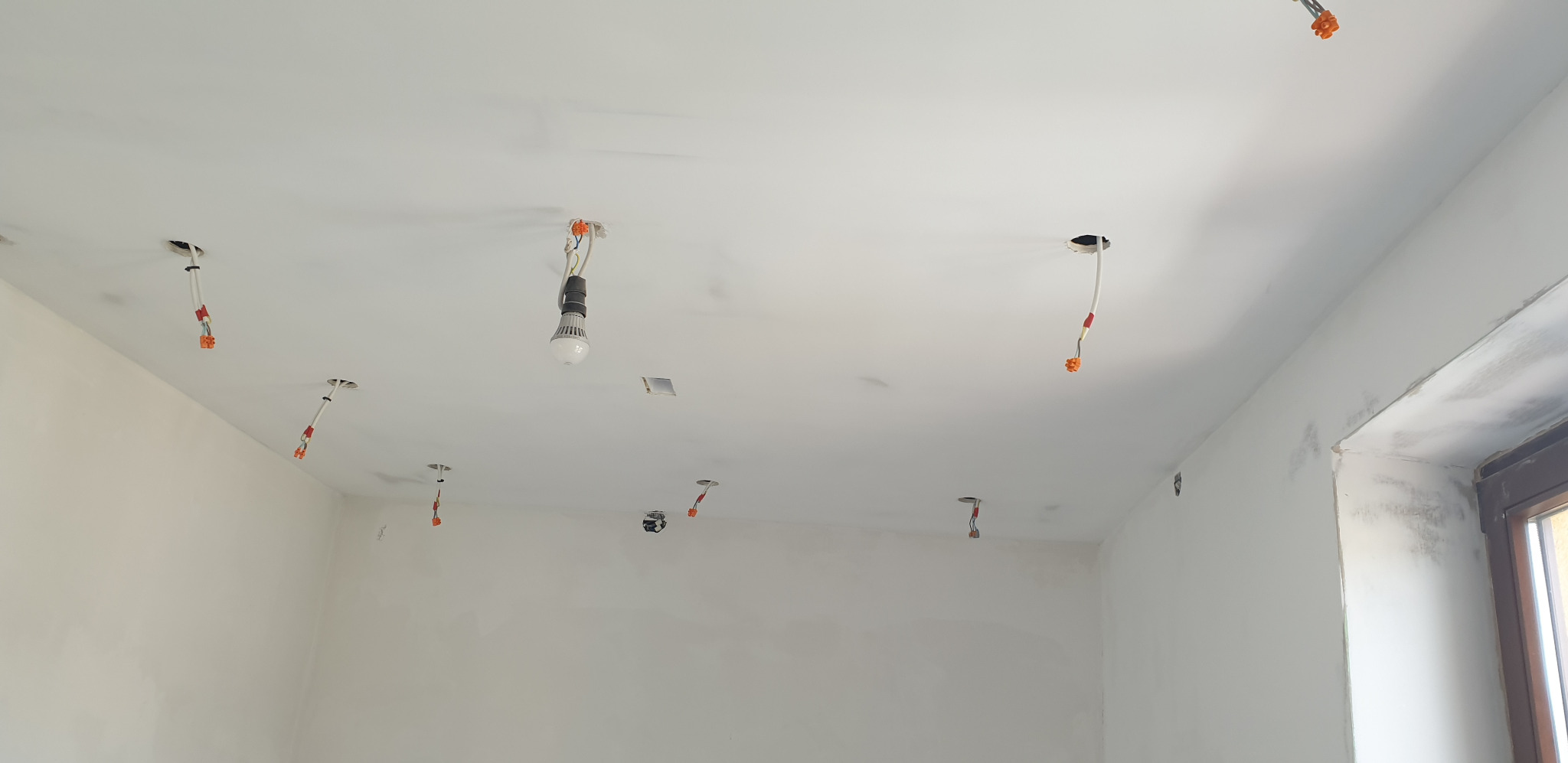 Wnętrze w trakcie remontu z widocznymi przewodami elektrycznymi wystającymi z sufitu, jeden z przewodów zakończony żarówką LED, ściany świeżo pomalowane na biało, widoczne ślady po szpachlowaniu