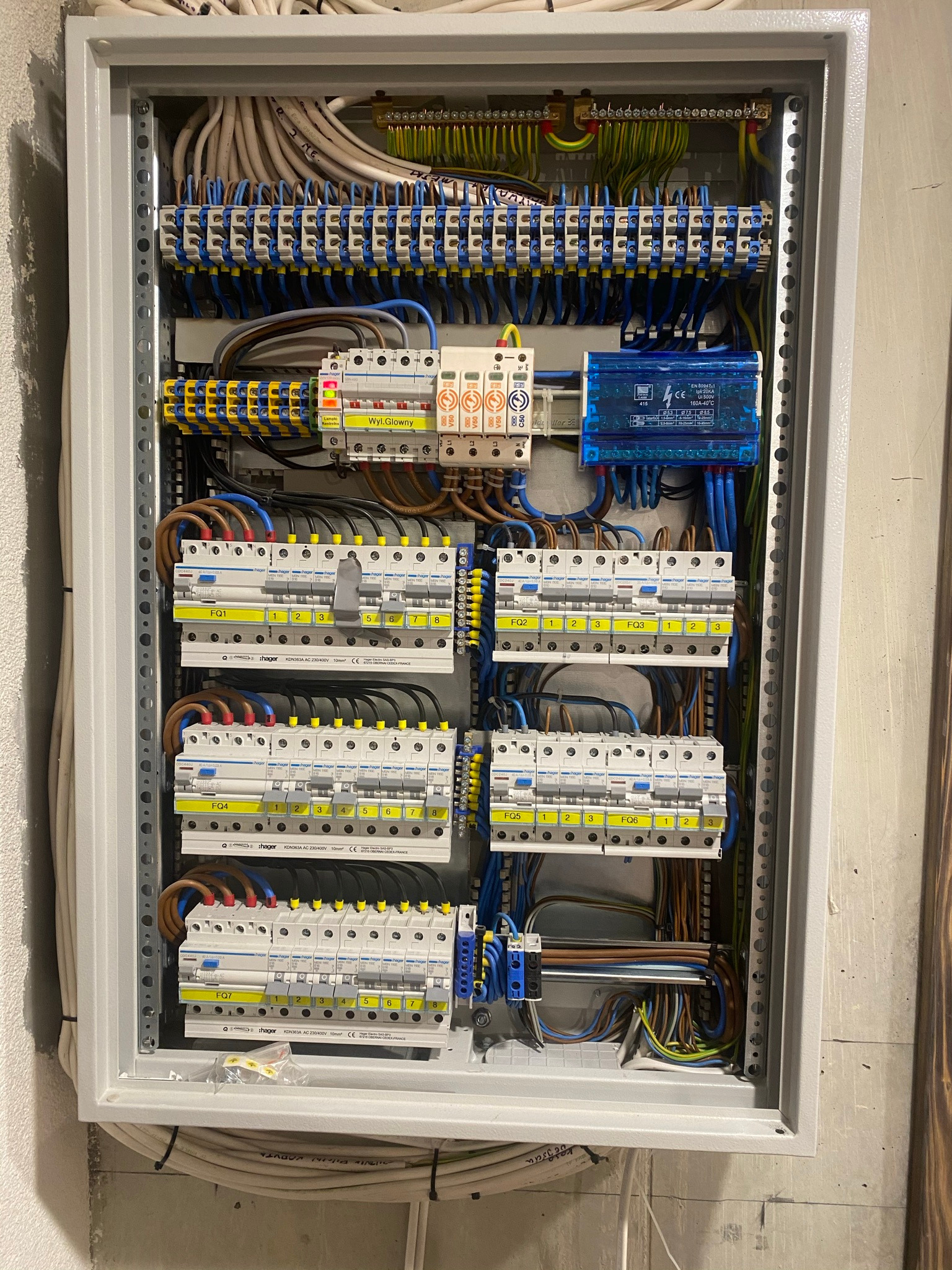 Rozdzielnica elektryczna w metalowej obudowie, widoczne liczne bezpieczniki Hager, przewody w różnych kolorach i niebieski moduł przeciwprzepięciowy, zamontowana na szarej ścianie.