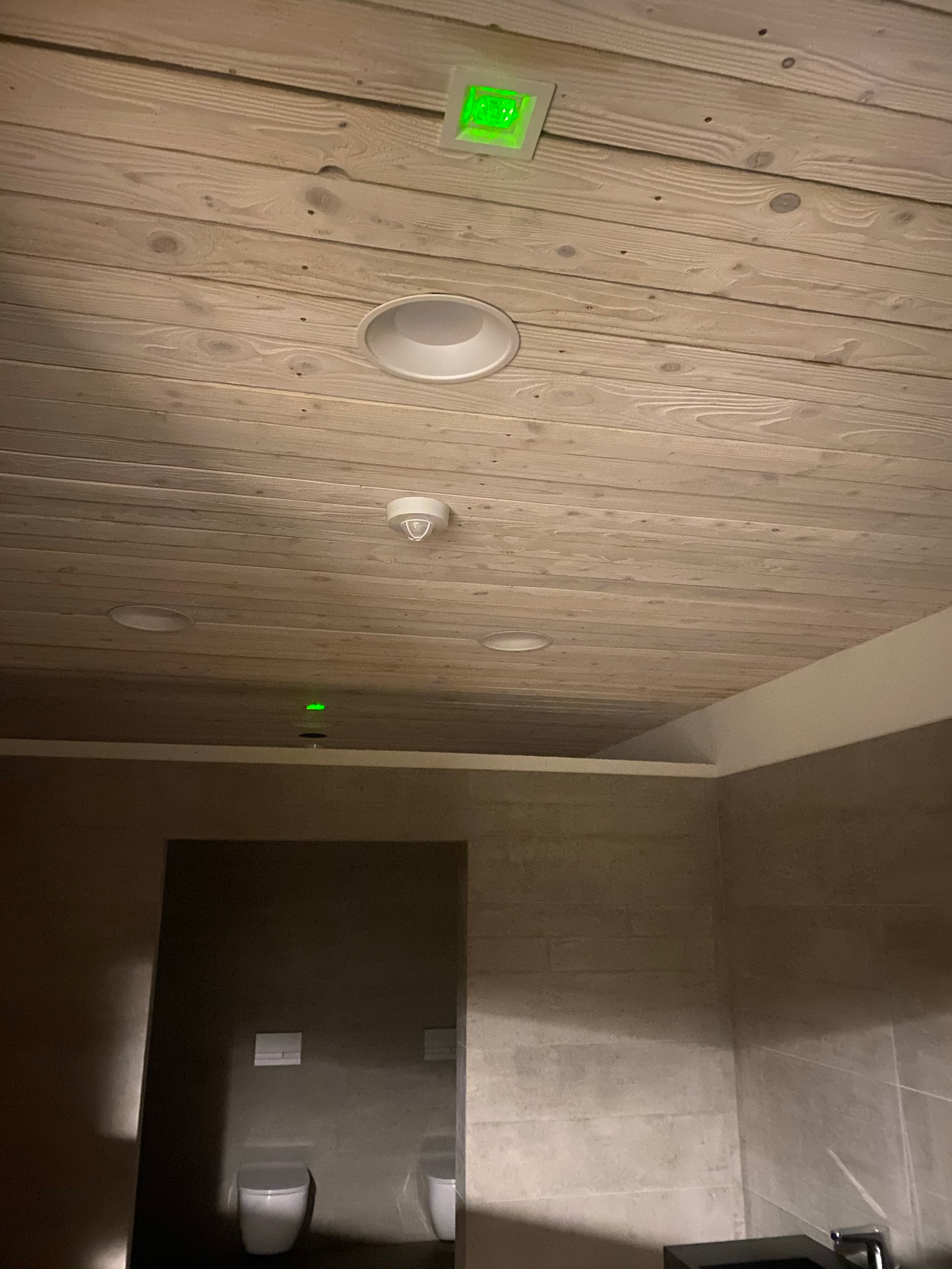 Drewniany sufit z zamontowanym oświetleniem LED, czujnikiem dymu i lampą sufitową w łazience z szarymi płytkami i podwieszaną toaletą.