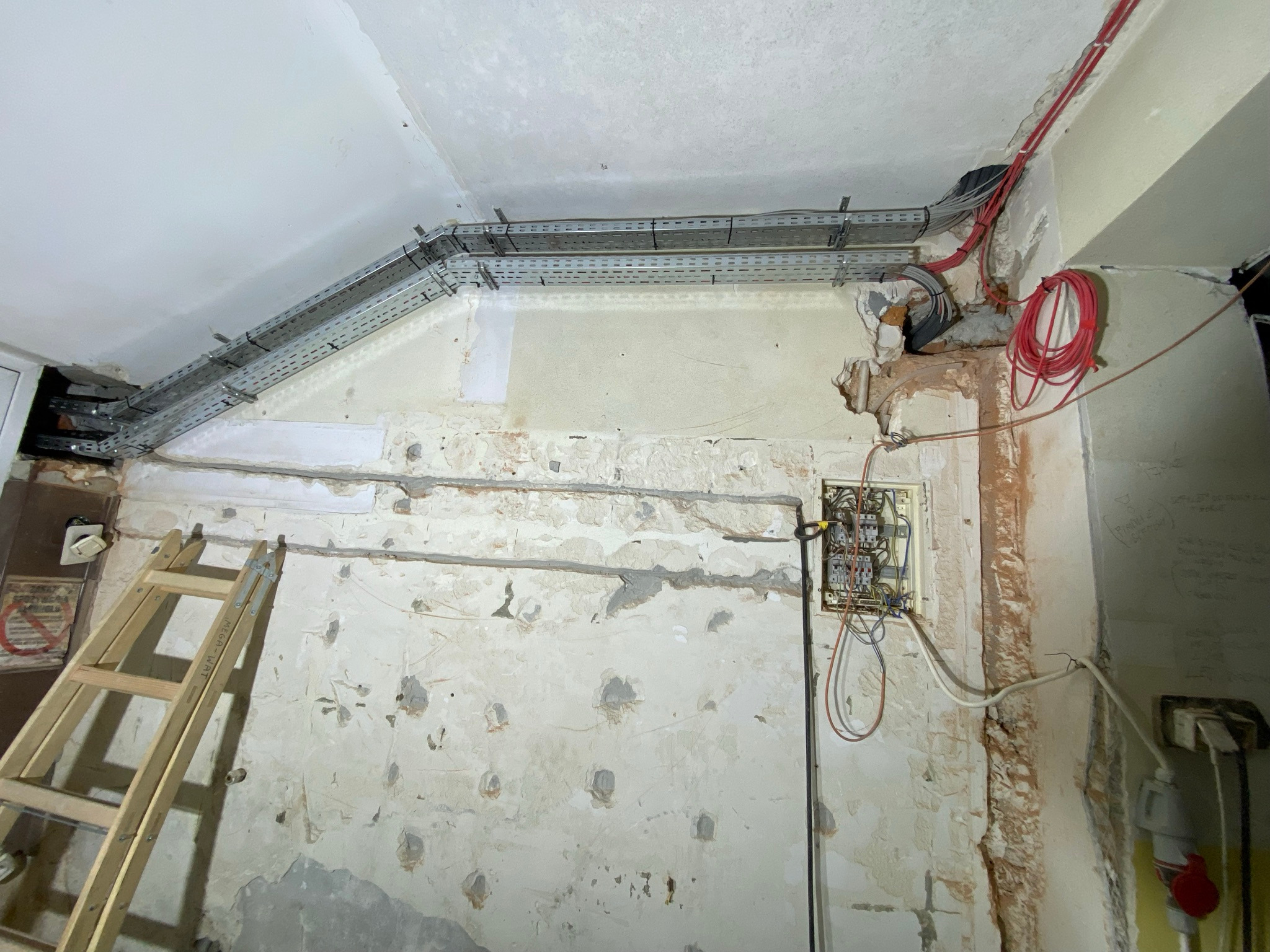Wnętrze w trakcie remontu z widoczną drabina MEGA-WAT, korytkami kablowymi pod sufitem, przewodami elektrycznymi na ścianie i skrzynką bezpiecznikową w zabudowie.