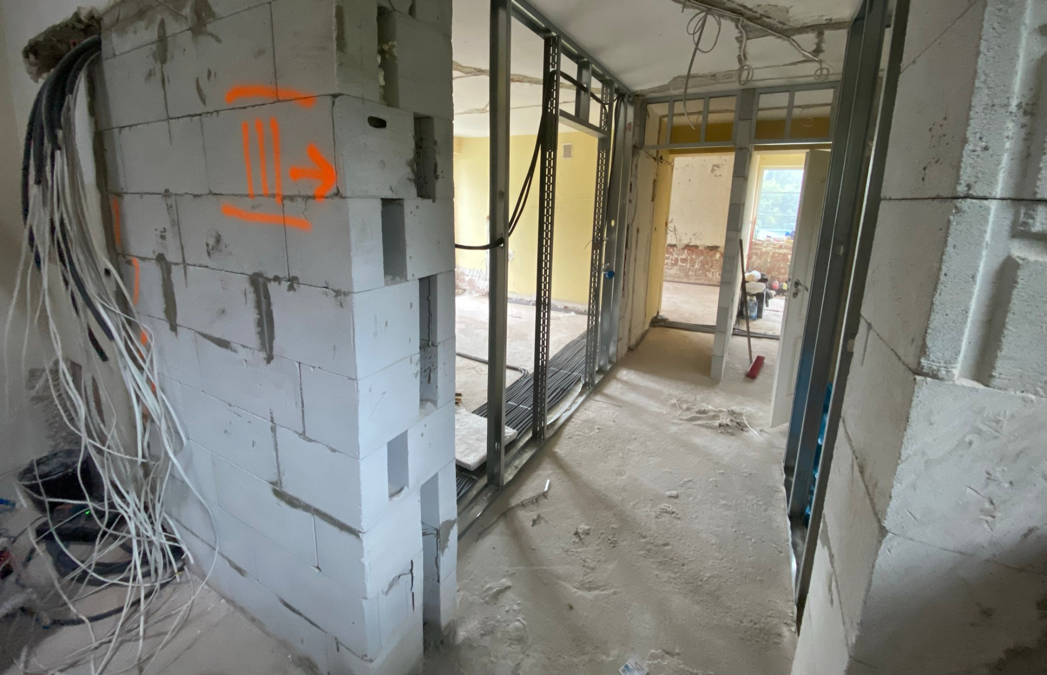 Wnętrze w trakcie remontu: murowana ściana z betonu komórkowego z pomarańczowymi oznaczeniami, metalowa konstrukcja ścianki działowej, przewody elektryczne zwisające ze ściany i sufitu, widok...