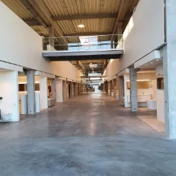 Długi korytarz w budynku komercyjnym z wyeksponowanymi stanowiskami łazienkowymi po bokach, betonowa podłoga i sufit z widoczną konstrukcją, oświetlenie punktowe, balustrada na antresoli.