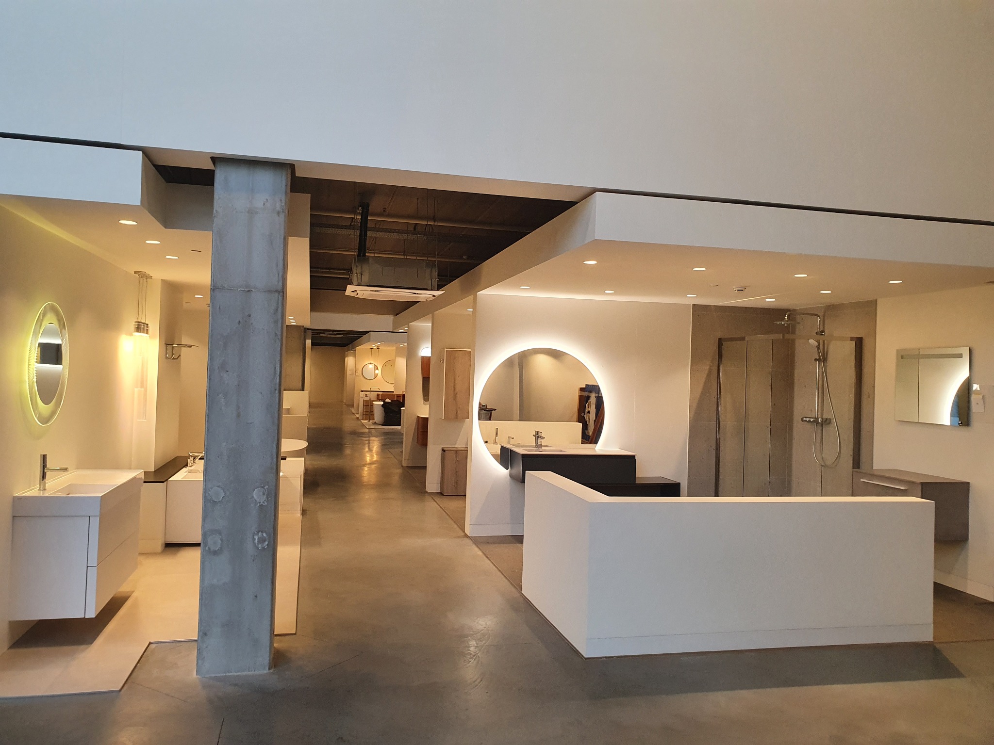Wnętrze salonu łazienek z ekspozycją umywalek z szafkami, kabiny prysznicowej i oświetlonych luster w okrągłych ramach, widok na przestrzeń z betonowym filarem.