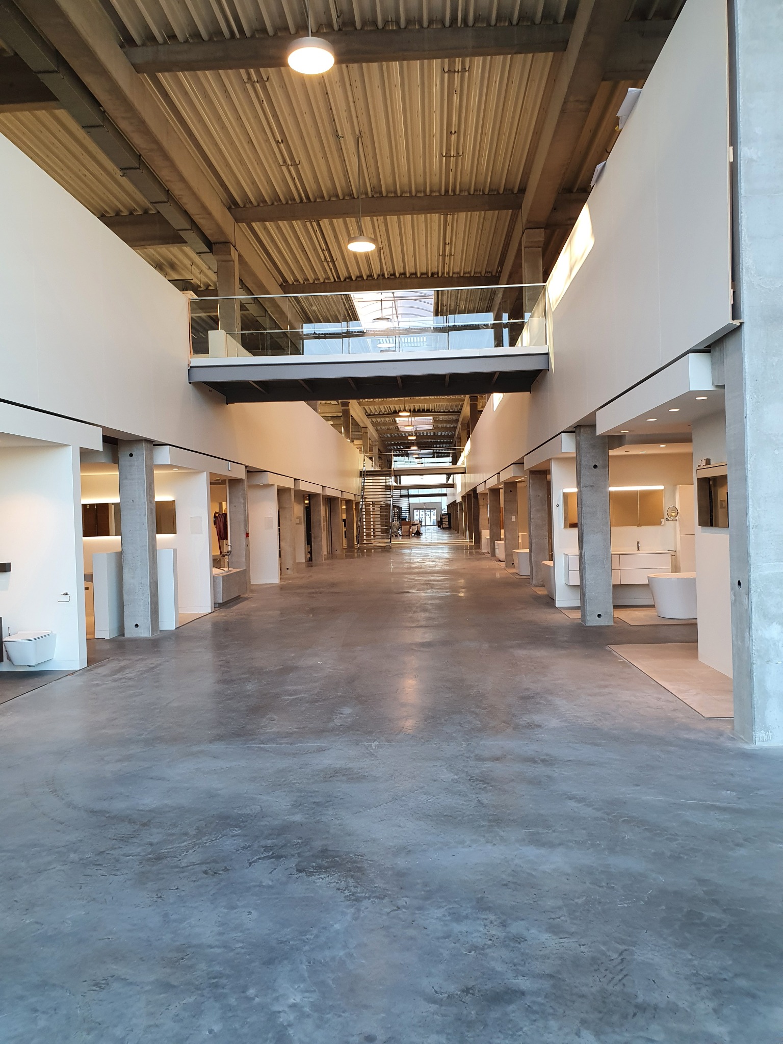 Długi korytarz w budynku komercyjnym z wyeksponowanymi stanowiskami łazienkowymi po bokach, betonowa podłoga i sufit z widoczną konstrukcją, oświetlenie punktowe, balustrada na antresoli.