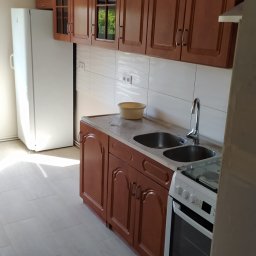 GS-Bud - Wykończona kuchnia z brązowymi szafkami z zaokrąglonymi drzwiczkami, białymi płytkami na ścianie, dwukomorowym zlewem i białą lodówką.