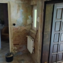 GS-Bud - Pokój w trakcie remontu: ściany z odsłoniętą cegłą i resztkami starej farby, grzejnik pod oknem, kubeł z zaprawą, narzędzia i zmiotka na podłodze z płytek.