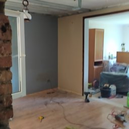 GS-Bud - Wnętrze w trakcie remontu: widoczne odsłonięte belki sufitowe, fragment ściany pomalowany na szaro, drewnopodobna podłoga, narzędzia rozrzucone na podłodze, oraz fragment muru z cegły po lewej...