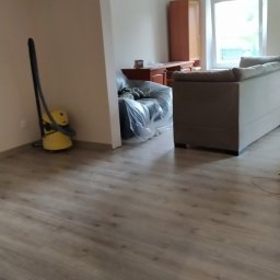 GS-Bud - Wnętrze pokoju po remoncie: widoczna nowa podłoga imitująca drewno, odkurzacz i meble przykryte folią ochronną.