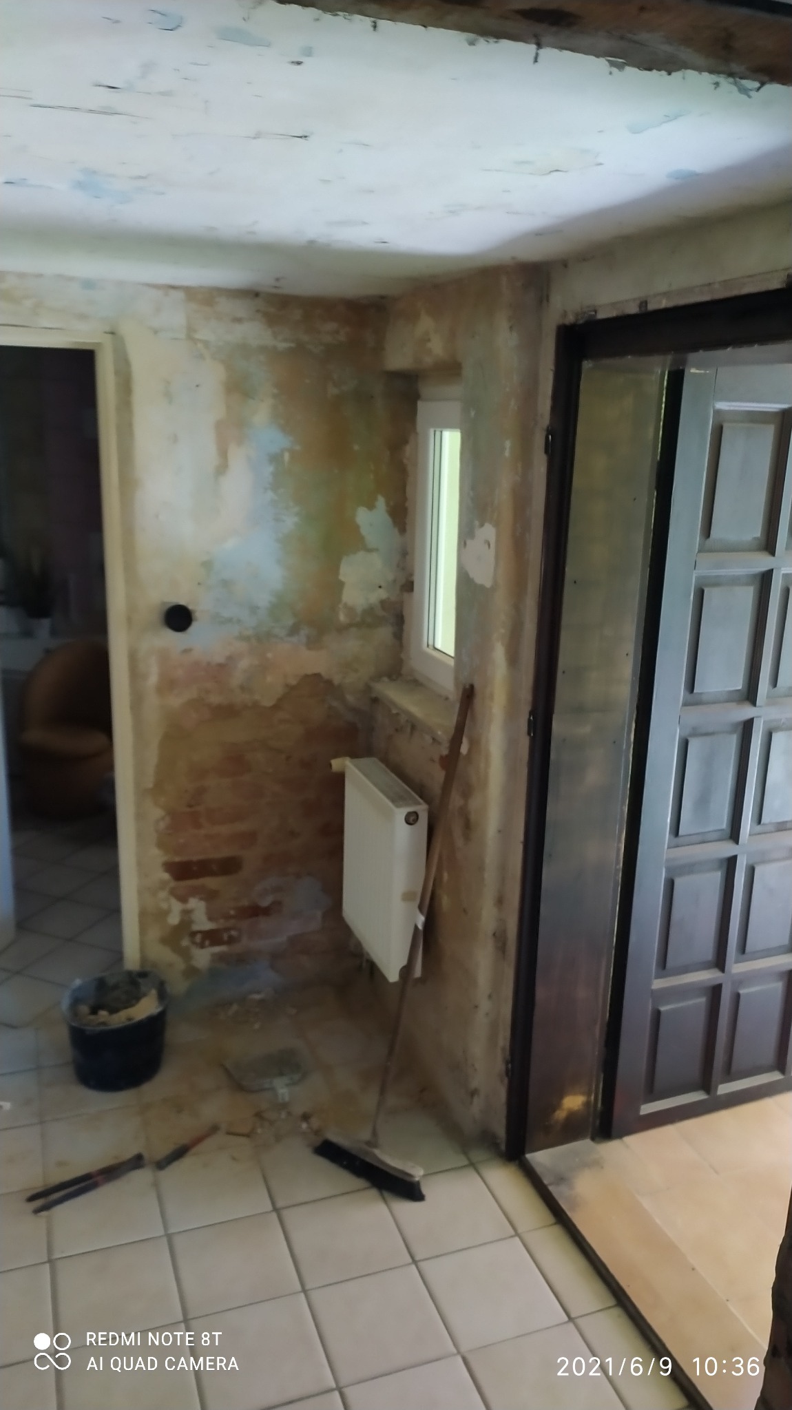 Pokój w trakcie remontu: ściany z odsłoniętą cegłą i resztkami starej farby, grzejnik pod oknem, kubeł z zaprawą, narzędzia i zmiotka na podłodze z płytek.