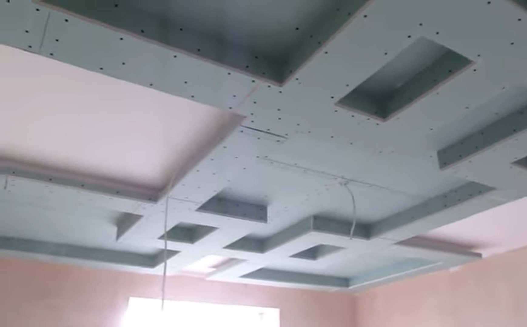 Sufit podwieszany z płyt kartonowo-gipsowych w trakcie montażu, z widocznymi otworami na śruby i przewodami elektrycznymi, tworzący geometryczny wzór z wgłębieniami.