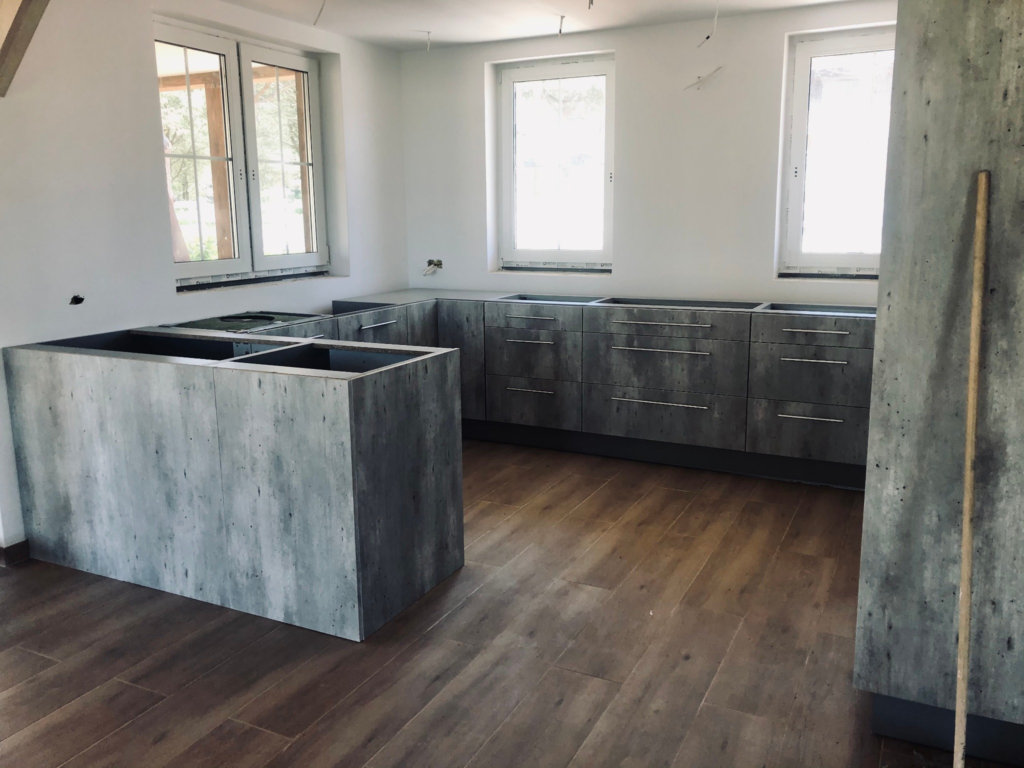 Surowa kuchnia z szarymi frontami i wyspą w stylu betonu, bez blatu, na tle jasnych ścian i drewnopodobnej podłogi w nowym domu.