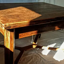 Kwadratowy, drewniany stół z wysuwanym blatem, oświetlony popołudniowym słońcem, rzucającym wyraźny cień na blat i podłogę.