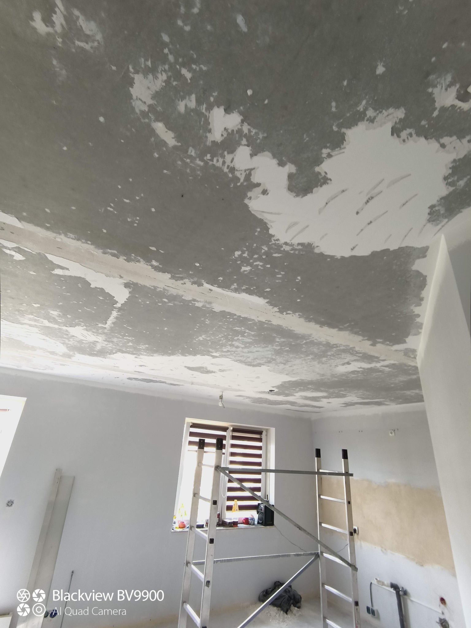 Pokój w trakcie przygotowania do malowania: sufit z częściowo usuniętą starą farbą, widoczna drabina aluminiowa, okno z żaluzjami i fragment ściany z niedokończonymi pracami.