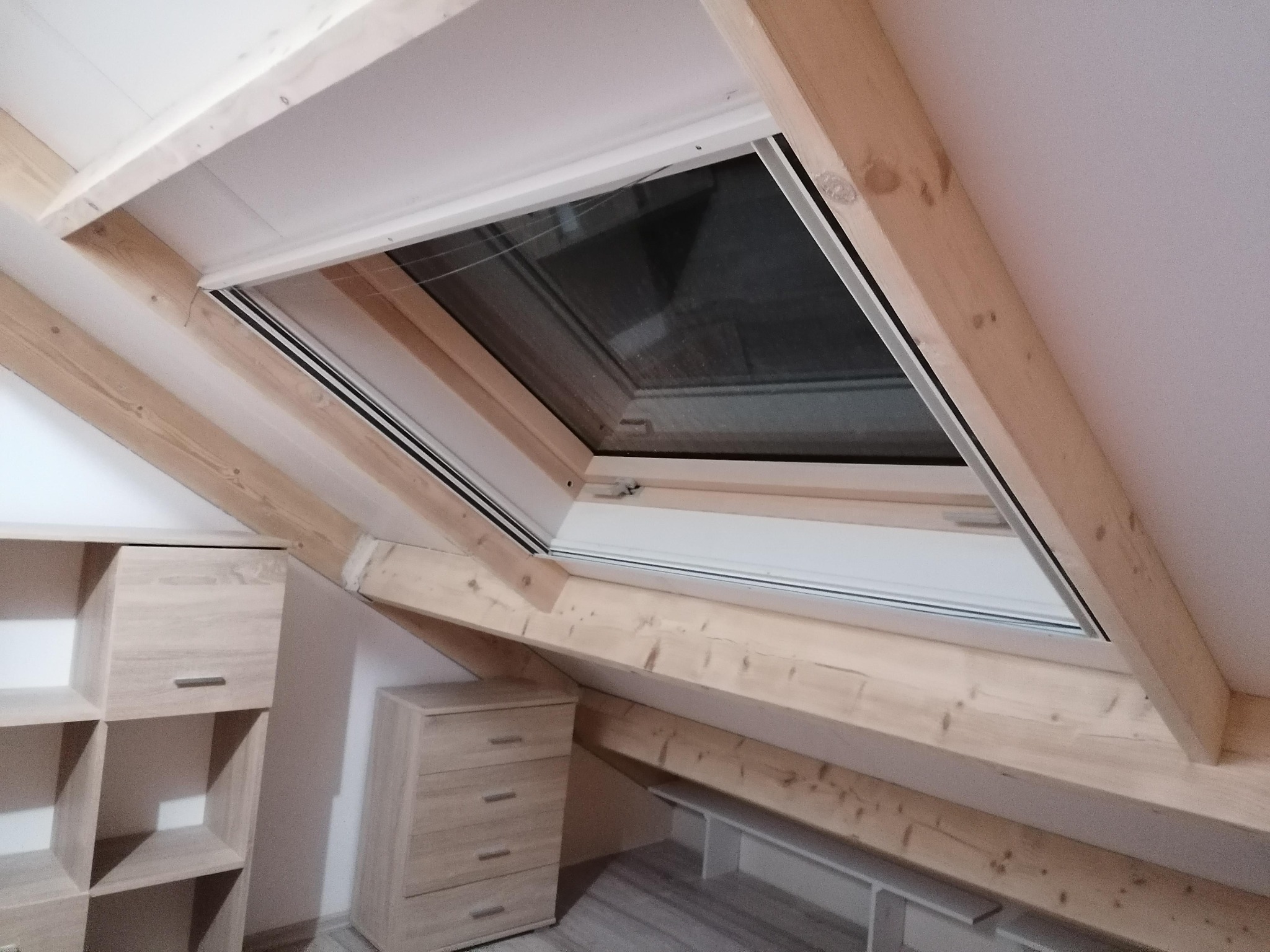 Poddasze z oknem dachowym, drewnianą konstrukcją nośną i zabudową meblową w jasnym odcieniu drewna.