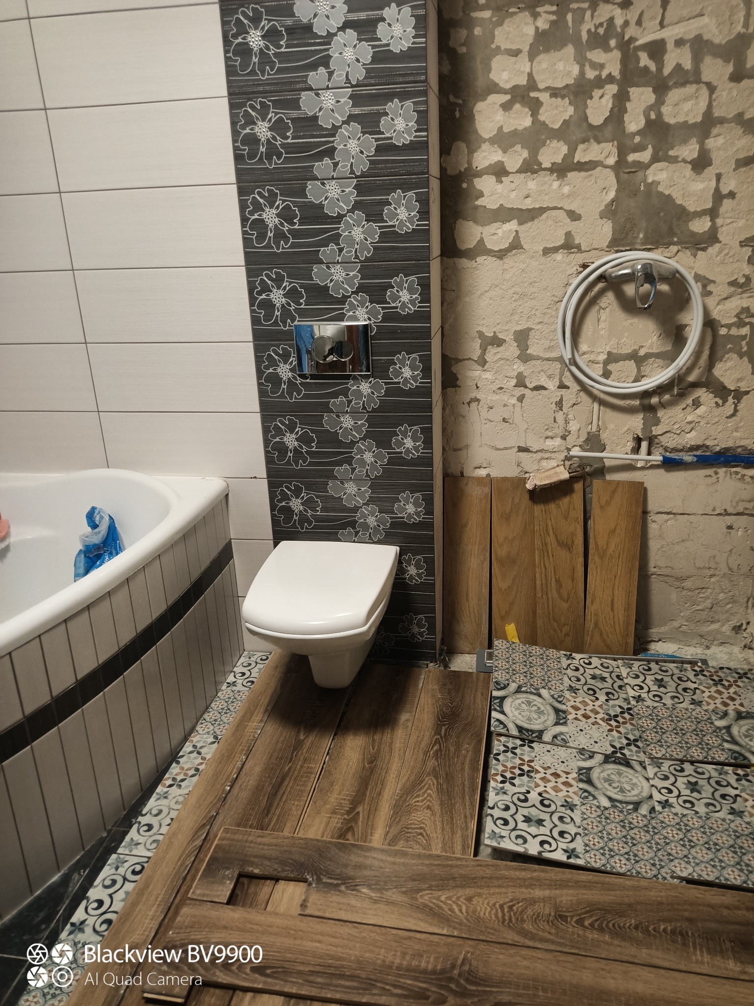 Łazienka w trakcie remontu: ułożone płytki podłogowe z różnymi wzorami, częściowo ułożone panele podłogowe, widoczne płytki ścienne z motywem kwiatowym oraz surowa ściana z instalacją wodną.