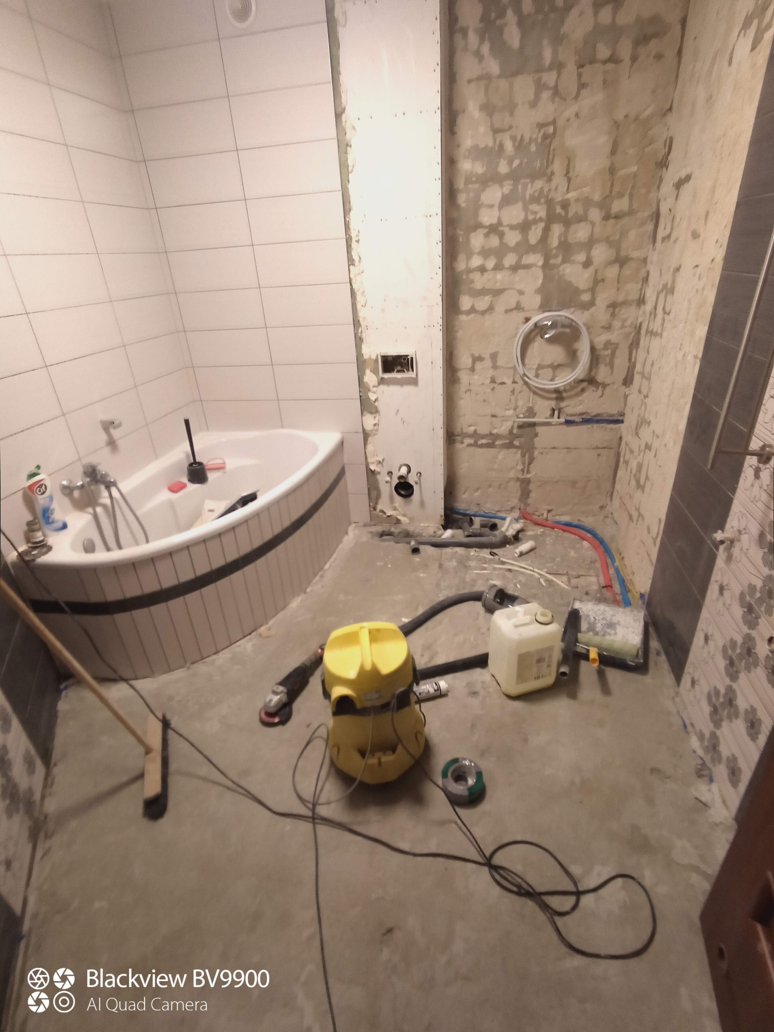 Łazienka w trakcie remontu: narożna wanna z biało-szarą obudową, częściowo położone białe płytki na ścianach, odsłonięta surowa ściana z instalacjami, żółty odkurzacz przemysłowy i narzędzia...