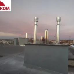 Instalacja wentylacyjna na dachu budynku z widocznymi kominami wentylacyjnymi, tło z zachodem słońca i panoramą miasta.