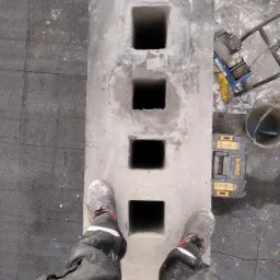 Betonowy moduł kominowy z czterema otworami, widok z góry, obok skrzynka narzędziowa DeWalt, w tle wciągarka linowa, robocze buty na module.