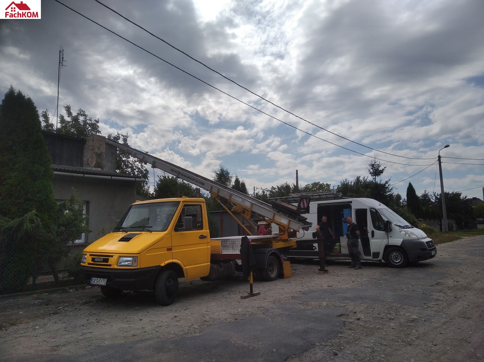Żółty samochód z wysuwaną drabiną pod oknem budynku, obok biały bus z otwartymi drzwiami i dwoma mężczyznami w środku, pochmurne niebo w tle.