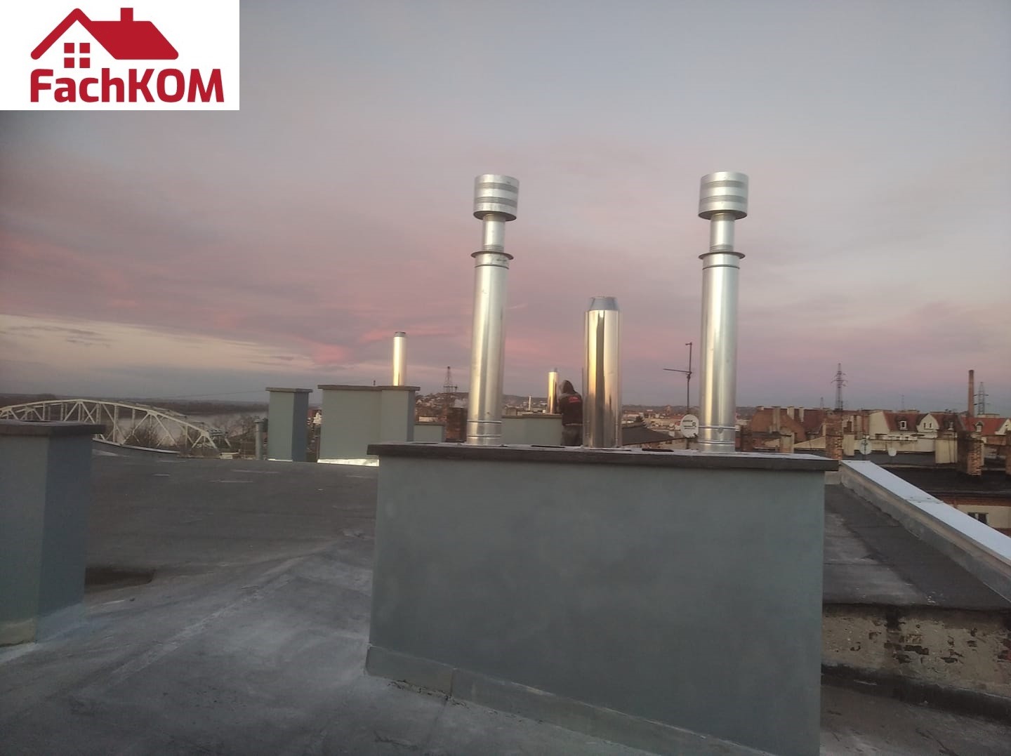 Instalacja wentylacyjna na dachu budynku z widocznymi kominami wentylacyjnymi, tło z zachodem słońca i panoramą miasta.