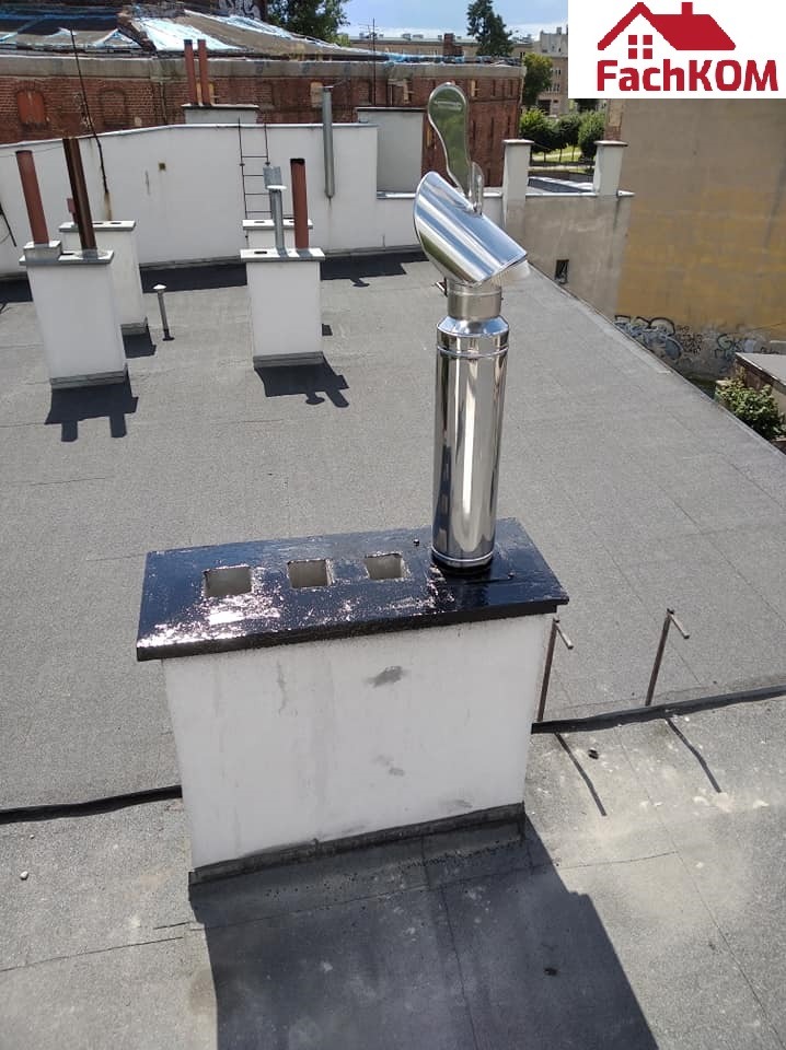 Błyszczący, chromowany wkład kominowy na dachu pokrytym papą, z widocznymi innymi kominami w tle.