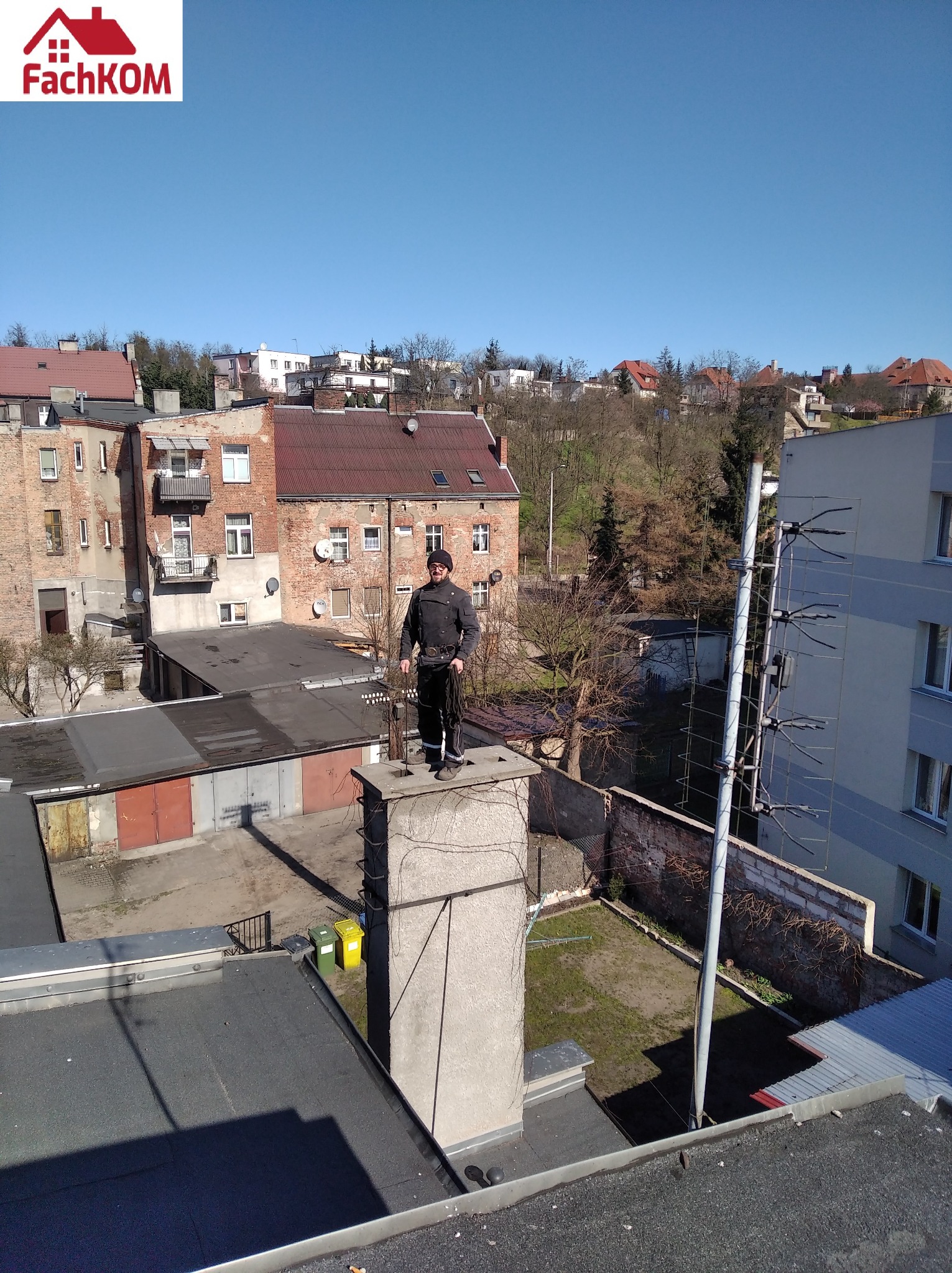 Kominiarz w tradycyjnym stroju stoi na szczycie komina z narzędziami, widok z góry na dachy budynków i podwórko w słoneczny dzień.