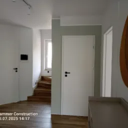 Nowoczesny przedpokój z drewnianymi schodami, białymi drzwiami i podłogą. Uwagę przykuwa duże, okrągłe lustro z drewnianą ramą. Minimalistyczne oświetlenie sufitowe.