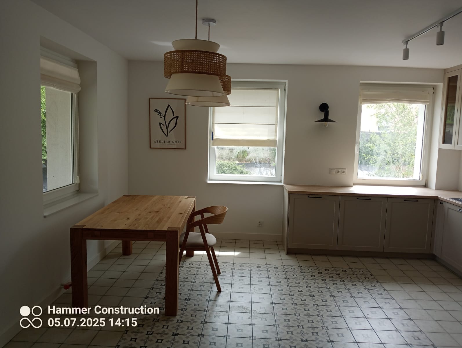 Jasna kuchnia z drewnianym stołem, krzesłem i wzorzystą podłogą. Okna z roletami rzymskimi wpuszczają naturalne światło. W tle szafki kuchenne i lampa wisząca.