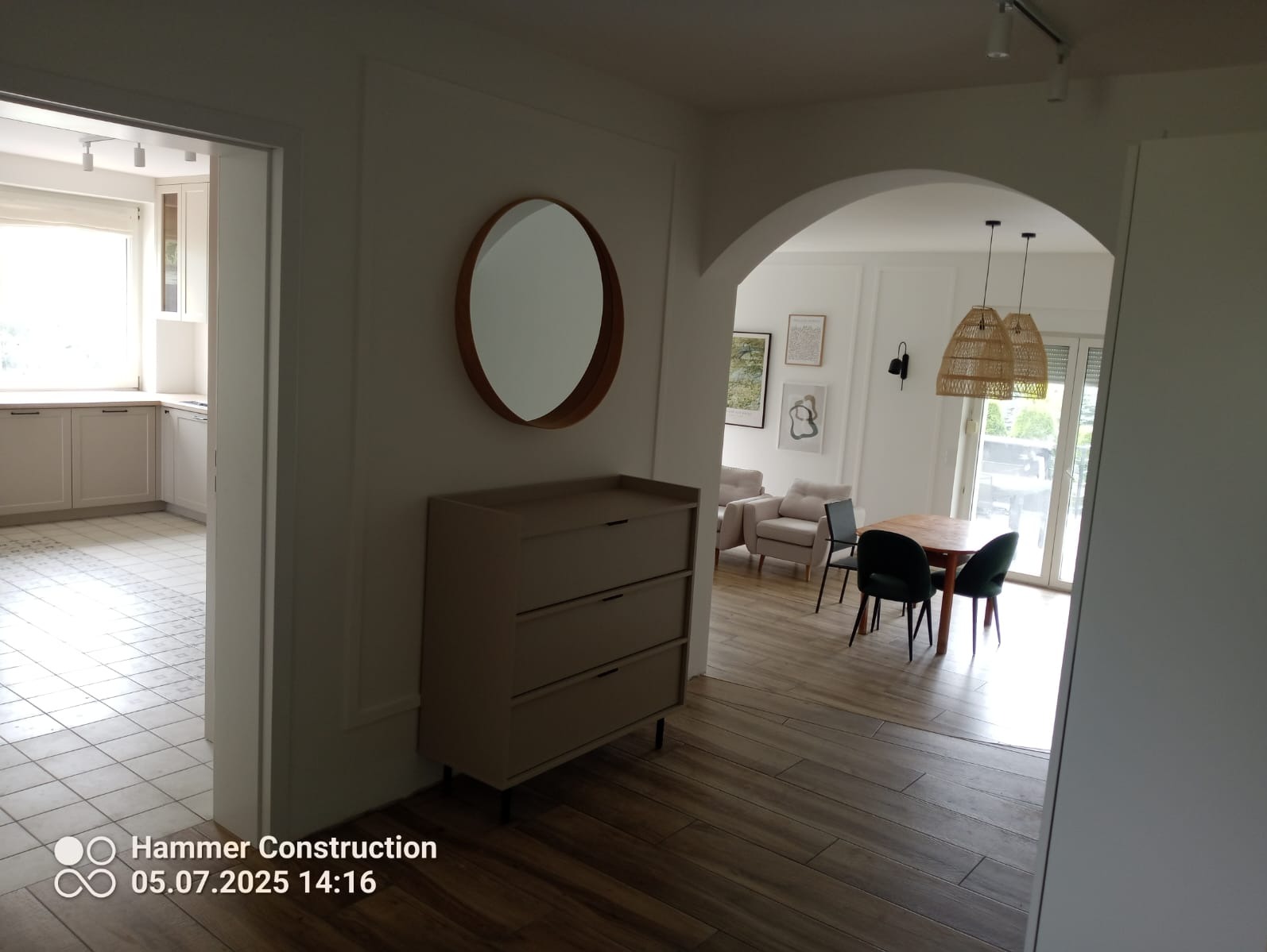 Jasne wnętrze po remoncie: widok na salon z okrągłym lustrem, komodą i kuchnią w tle. Parkiet ułożony w jodełkę, białe ściany z dekoracyjnymi listwami.