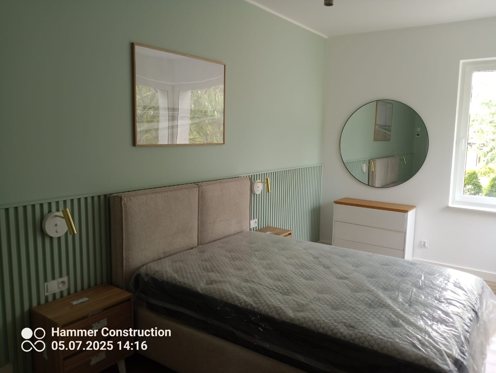 Pokój po remoncie: łóżko z materacem w folii, szafka nocna, komoda, okrągłe lustro, obraz w ramie, lampki ścienne i zielono-białe panele dekoracyjne na ścianie.